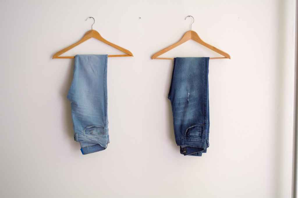 Two pairs of jeans hanging on two wooden hangers, one light blue, one dark blue. On a white background