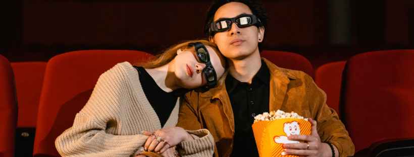 Man and woman sitting in an empty cinema with a yellow box of popcorn and 3D glasses on. The woman is resting her head on the mans right shoulder.